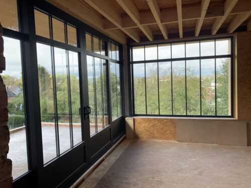 Intérieur d'une véranda avec structure en bois et baies vitrées noires donnant sur une terrasse