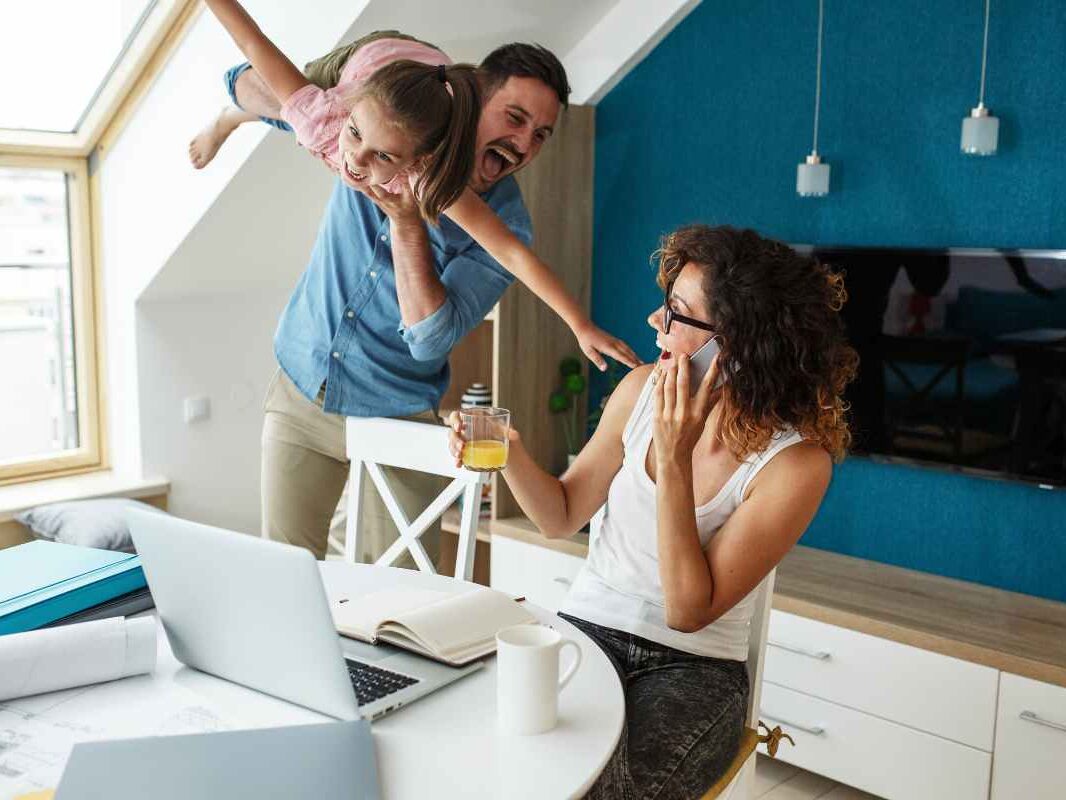 Papa qui fait l'avion avec sa fille et rigole avec sa femme assise à une table devant un ordinateur