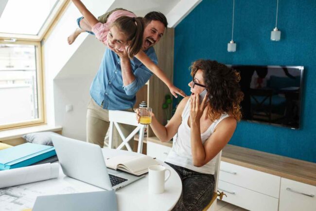 Papa qui fait l'avion avec sa fille et rigole avec sa femme assise à une table devant un ordinateur