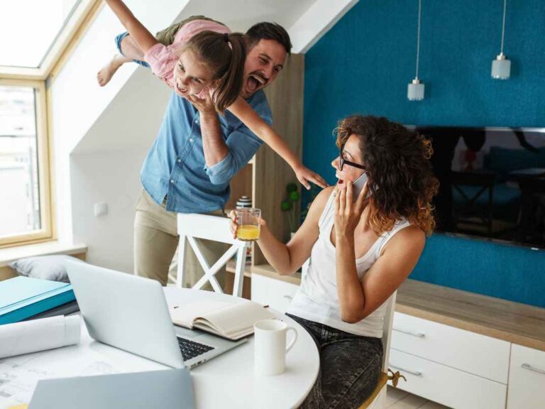 Papa qui fait l'avion avec sa fille et rigole avec sa femme assise à une table devant un ordinateur