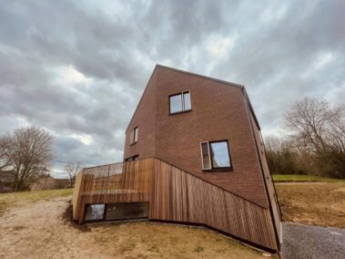 Maison en brique rouge avec bardage en bois avec trois fenêtres avec une partie du chassis en bois.