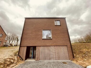 Maison en brique rouge avec un bardage en bois et deux fenêtres avec une partie du chassis en bois