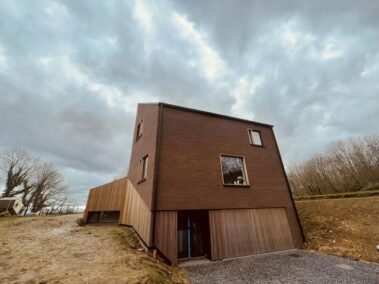 Maison en brique rouge avec un bardage en bois clair débordant sur la terrasse à gauche.